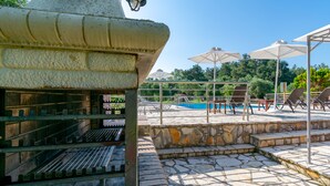 Un área junto a la piscina con un patio de baldosas de piedra, sillas de descanso, sombrillas y una parrilla.