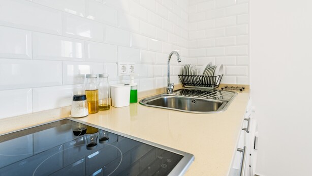 A modern kitchen with white tiles, a stainless steel sink, a dish rack, and a built-in induction cooktop.