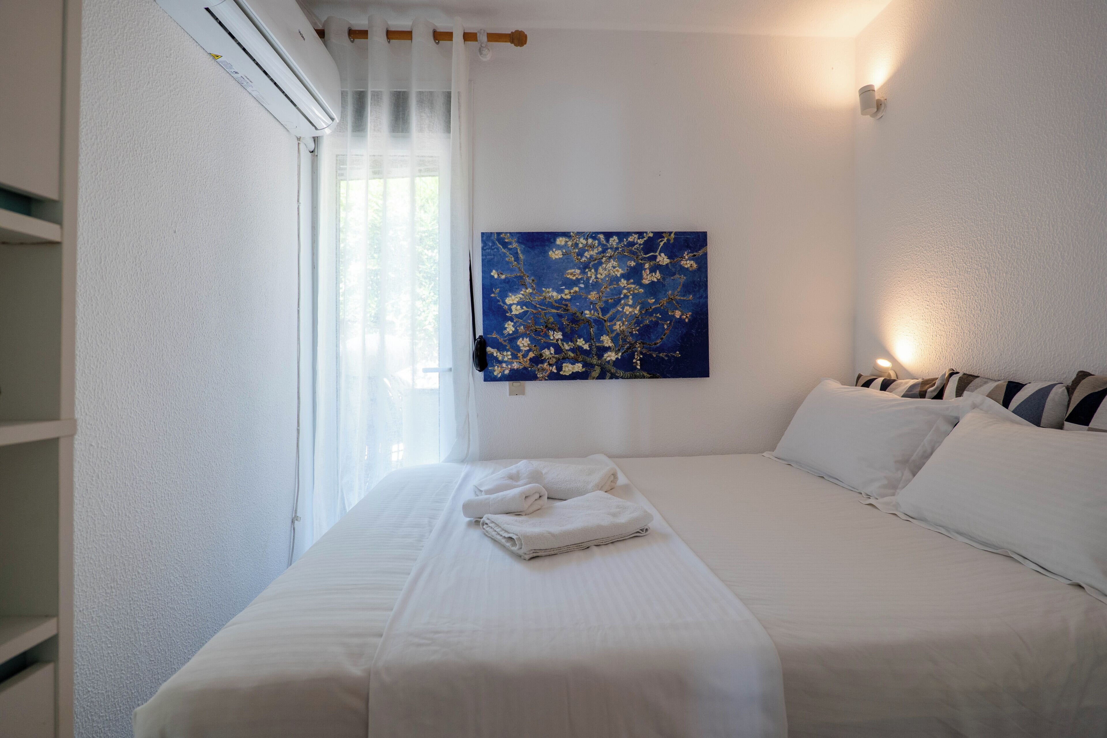 A hotel room with a bed, white curtains, an air conditioning unit, a painting on the wall, and a bedside lamp.