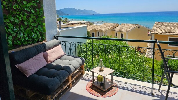 A balcony with a cushioned seating area, a small table with a lamp, and a view of the sea and buildings.