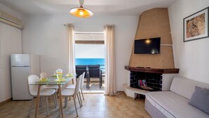 Uma sala de estar com lareira, televisão fixada na parede, espaço de refeições com cadeiras e mesa, e vista para o mar pela janela.