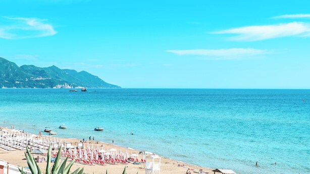 Uma praia com fileiras de espreguiçadeiras e guarda-sóis, mar azul cristalino e montanhas ao fundo.
