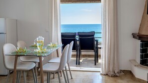 Uma área de refeições com mesa de vidro, cadeiras e vista para o mar através de uma porta.