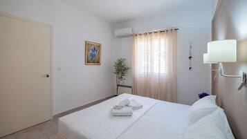 A hotel room with a bed, a door, an air conditioning unit, a window with curtains, a wall-mounted lamp, and a framed picture.