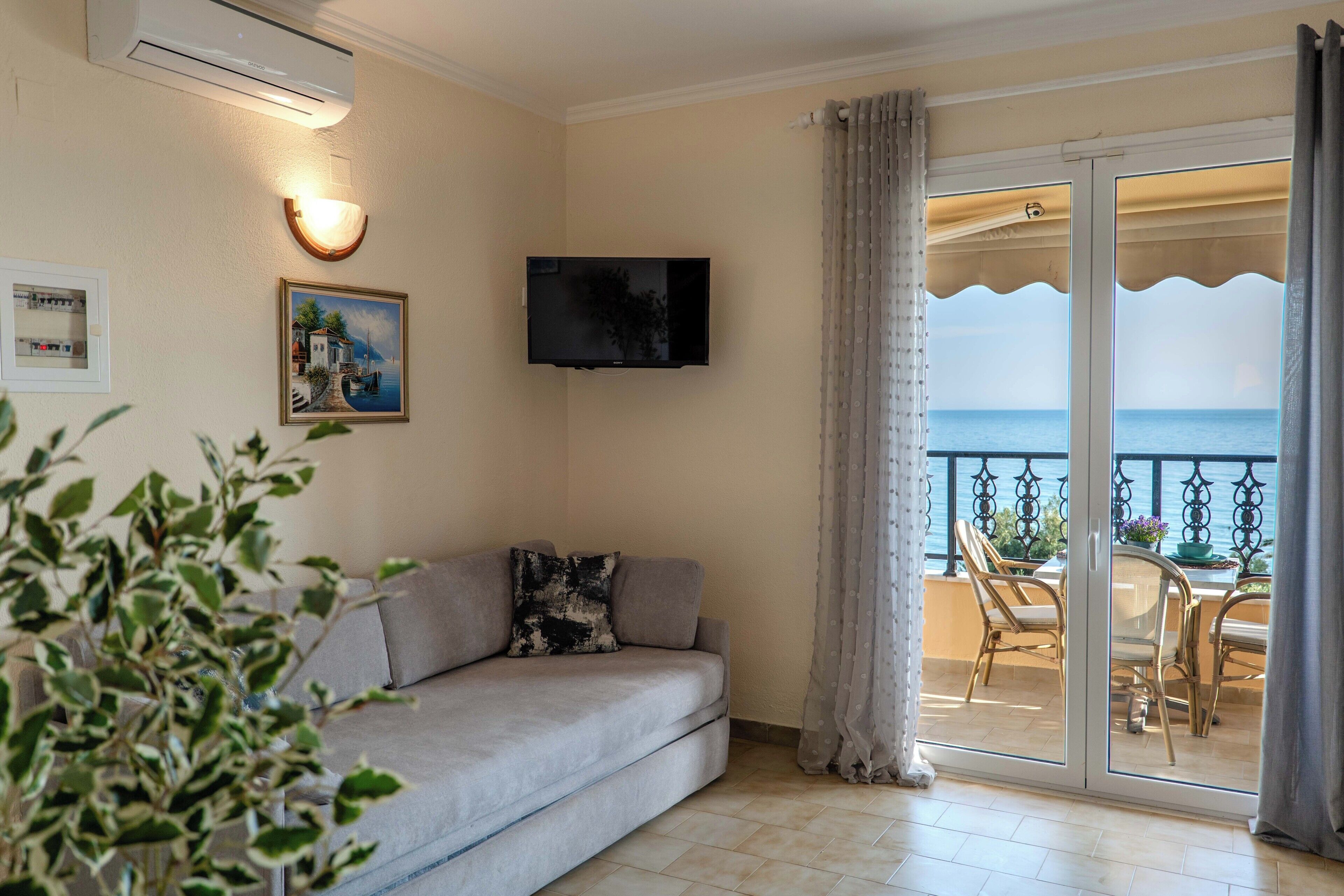 A living room with a sofa, a flat-screen TV, a painting, and a balcony with a view of the sea.