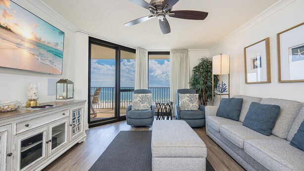 Een ruime woonkamer met een plafondventilator, een groot raam met uitzicht op het strand, een grijze bank, twee fauteuils en een witte kast.