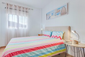 Ein Schlafzimmer mit gestreifter Bettdecke, einem Nachttisch mit Lampe, einem gerahmten Bild an der Wand und einem Fenster mit durchsichtigen Vorhängen.