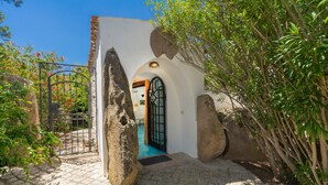 A stone pathway leading to a white building with a black gate, surrounded by greenery and large rocks. - Dream Villa with Terrace, Wi-Fi, Air Conditioning and Shared Pool (Porto Rotondo)