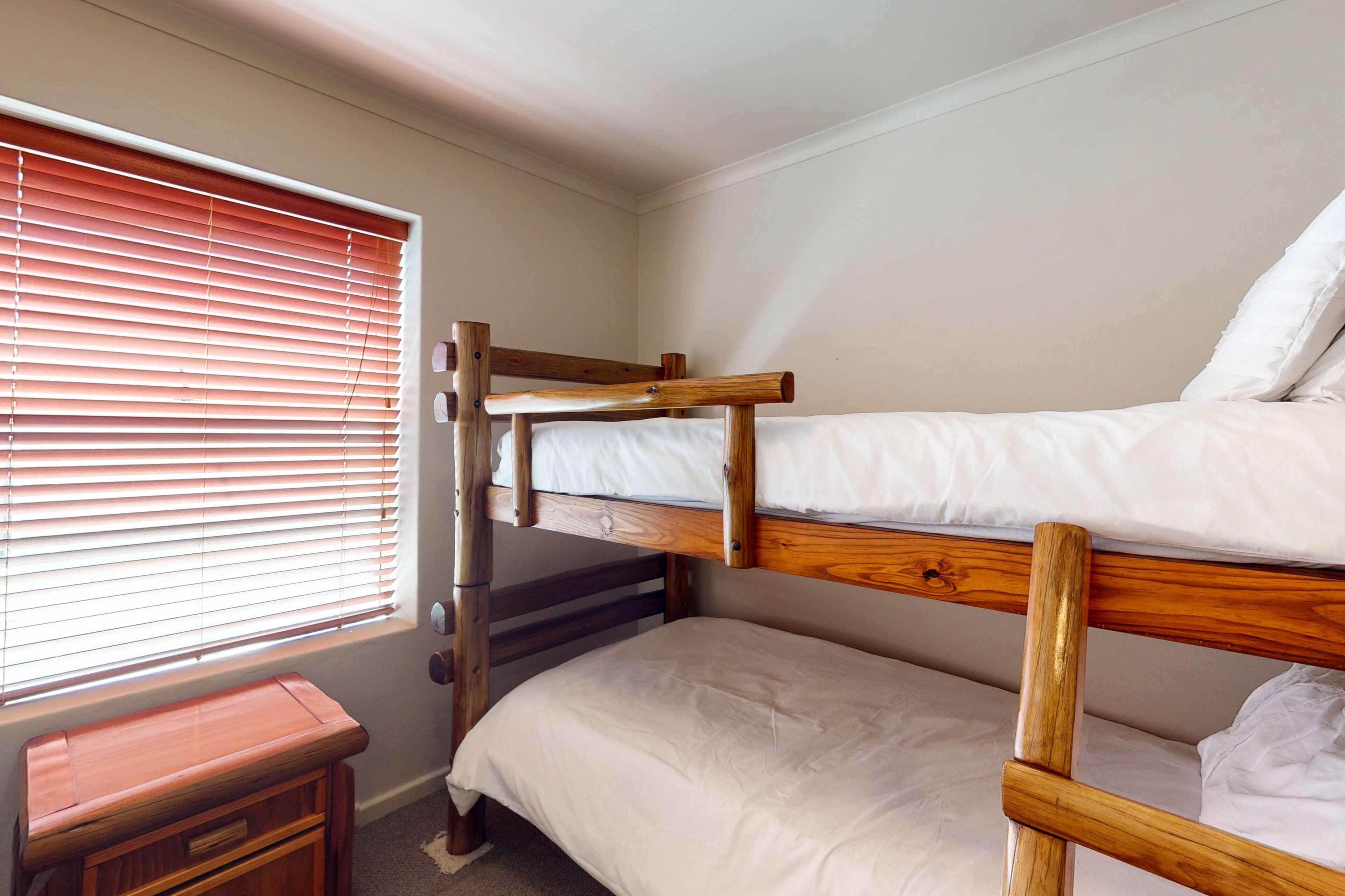 A bunk bed room with a wooden headboard, a small wooden nightstand, and a window with blinds.