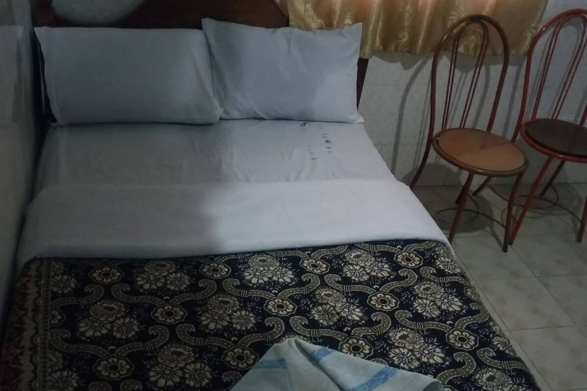 Una cama con sábanas blancas, una colcha estampada y un bolso en el suelo.