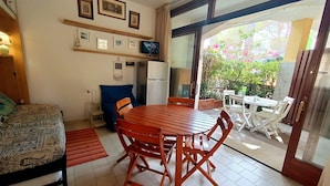 Una habitación con cama, una mesa de comedor con sillas, una pequeña cocina y un ventanal corredero que da a un área exterior con mesa y sillas.