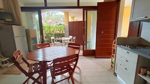 Una habitación con una mesa de comedor, sillas, una zona de cocina y un ventanal corredizo que da a un espacio exterior con plantas.