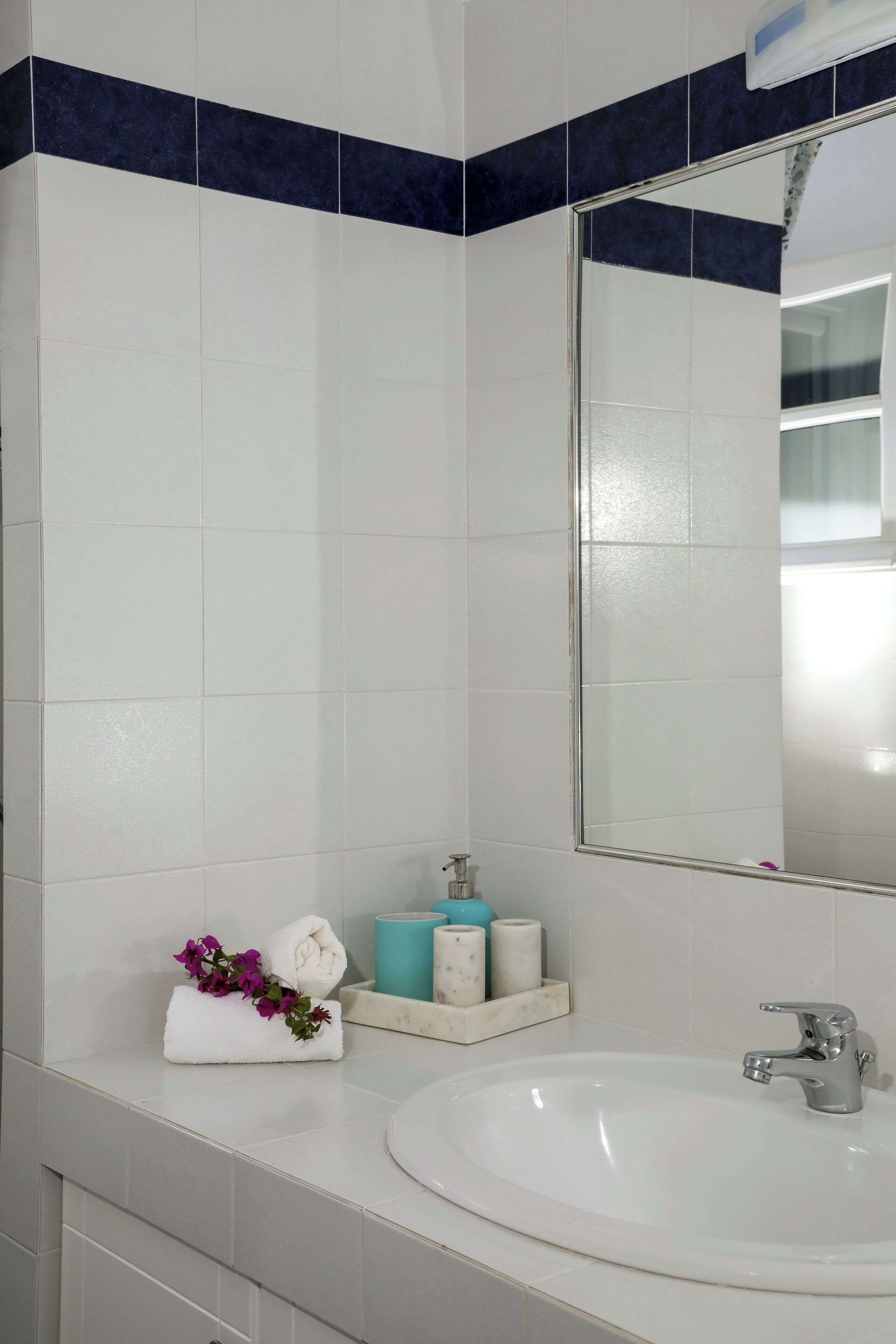 Casa de banho com azulejos brancos, um espelho grande, uma pia branca e uma toalha com flores no balcão.