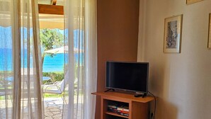 Quarto com televisão sobre suporte de madeira, mesa de vidro e uma cadeira. Há cortinas e vista para o exterior.