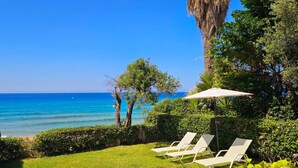 Um jardim com vista para o mar, duas espreguiçadeiras e um grande guarda-sol.