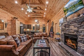 A log cabin interior with a fireplace, leather sofas, a dining area, and a kitchen.