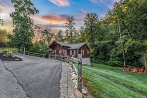 A cabin with a porch, surrounded by a forested area and a paved driveway. - Creekside On The Bluff 2 Bedroom Cabin by RedAwning (Sevierville)