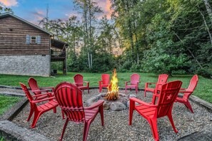 A fire pit with red chairs arranged around it, set in a gravel area with a wooden deck and a building in the background. - Creekside On The Bluff 2 Bedroom Cabin by RedAwning (Sevierville)
