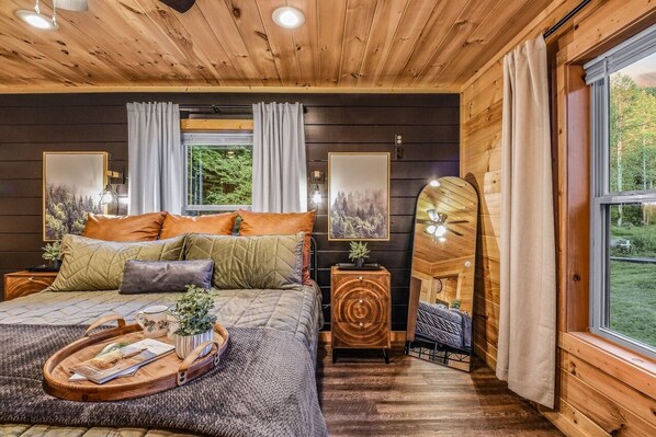 A cozy wooden cabin bedroom with a bed, curtains, a side table, and a window with a view of greenery.