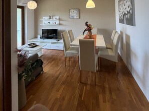 A modern living room with a dining area, wooden flooring, a flat-screen TV, white furniture, and wall-mounted shelves.