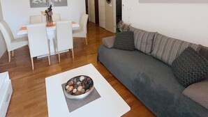 Een woonkamer met een grijze bank, een witte salontafel met een decoratieve schaal, witte stoelen en een eettafel met een bloevas.