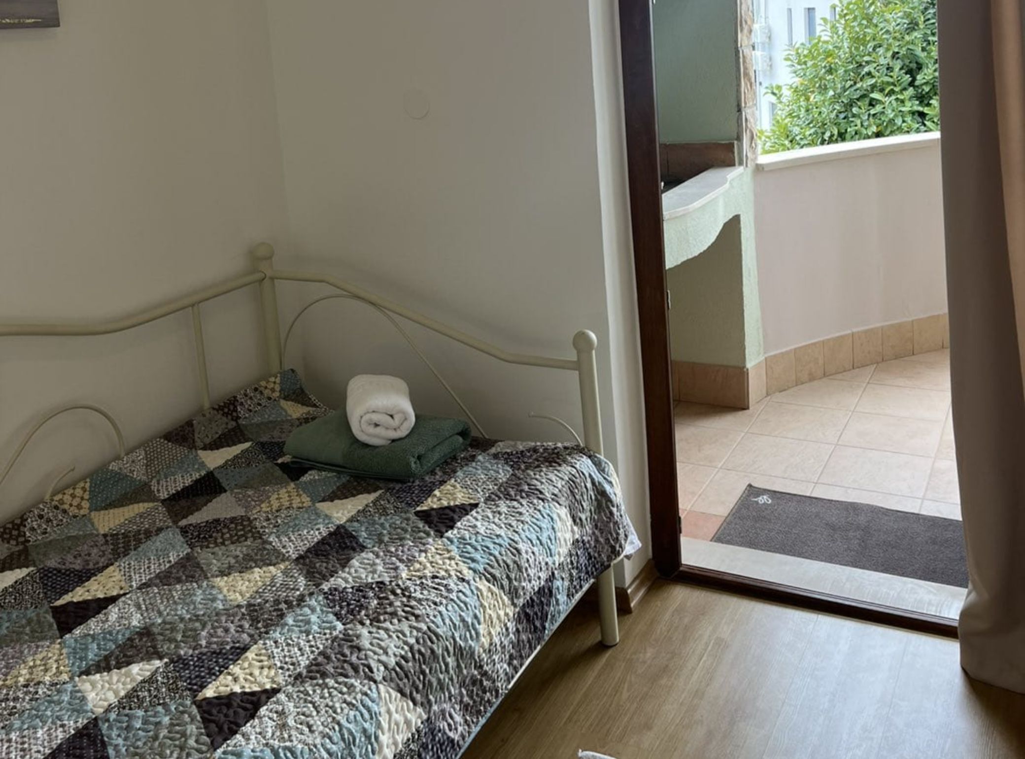 A small, single-bed room with a patterned quilt, a rolled towel, and a view of a balcony with a door leading outside.