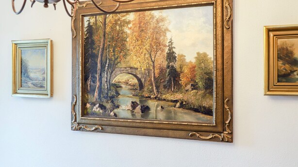 Pintura emoldurada de paisagem com uma ponte, rodeada por árvores e um corpo d'água.