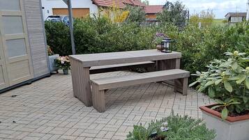 A wooden outdoor bench with a table, surrounded by potted plants and a paved patio.