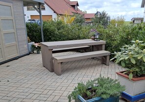 Un banc en bois installé à l’extérieur, avec une table, entouré de plantes en pot et bordé d’une terrasse dallée.