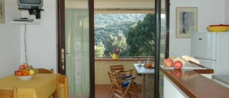 Una habitación con una mesa de comedor, sillas, un televisor, una encimera de cocina y un ventanal corredero que da a un balcón con vistas a una zona boscosa.