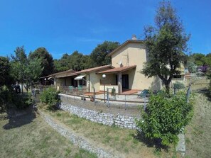 Une maison à deux étages, dotée d’un mur de soutènement en pierre, d’un patio couvert et d’un arbre au premier plan.