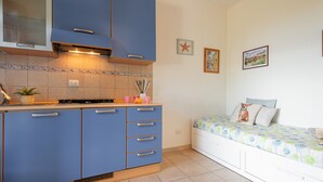 A kitchen with blue cabinets, a stove, and a sink. There is a bed with a floral bedspread and a pillow with a dog image. - Well-kept and with great views - Appartamenti Le Querce - Melograno (Capoliveri)