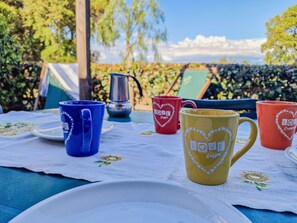 A table with coffee mugs and a kettle, set outdoors with a view of trees and a clear sky.