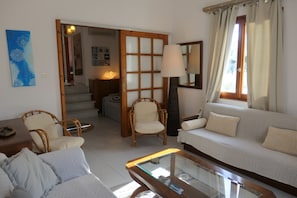 A living room with a sofa, chairs, a glass coffee table, a lamp, and a wooden door leading to another room.