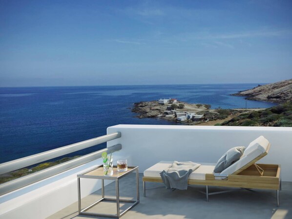 A balcony with a view of the sea, a small table with a drink, and a lounge chair.