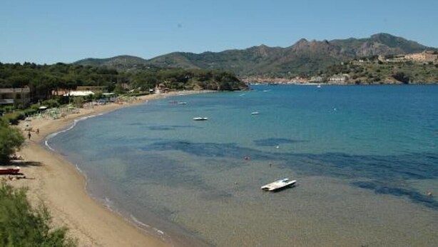 Una playa costera con agua cristalina, barcos y una montaña al fondo.
