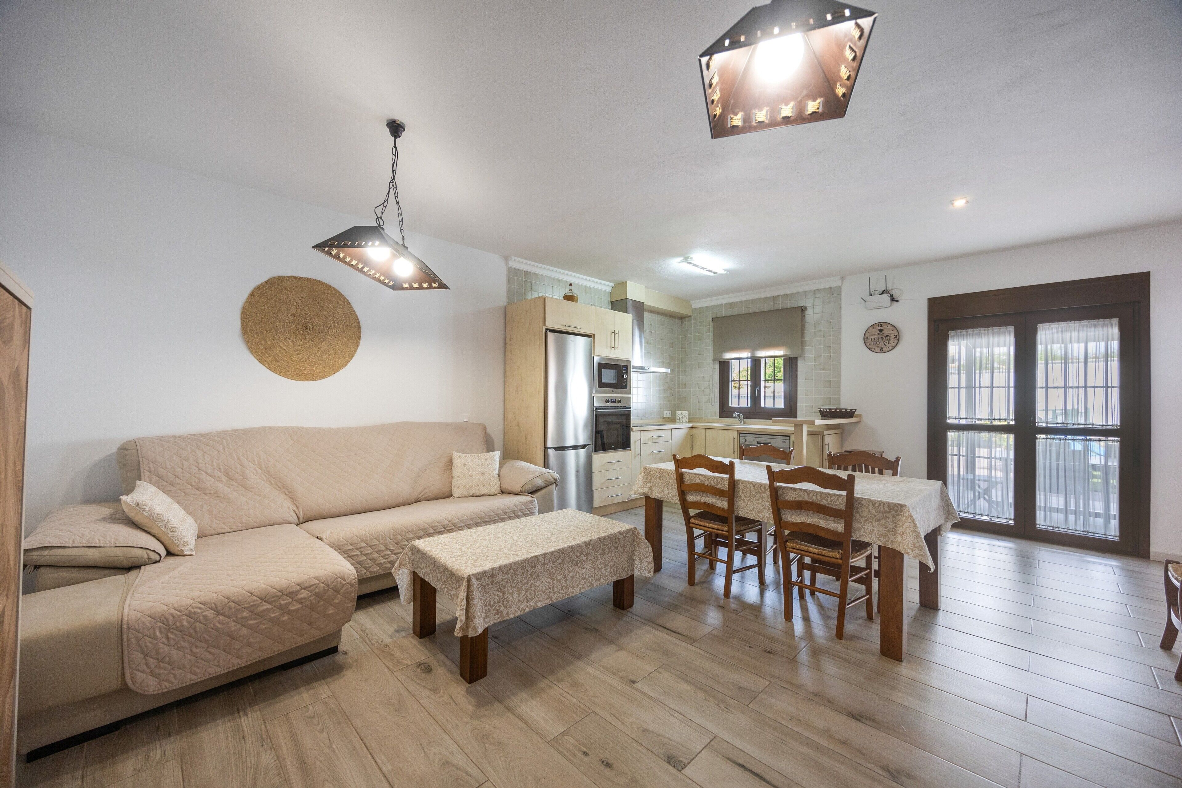 Een moderne woonkamer met een bank, eettafel en stoelen, een keuken met apparatuur, en een groot raam.