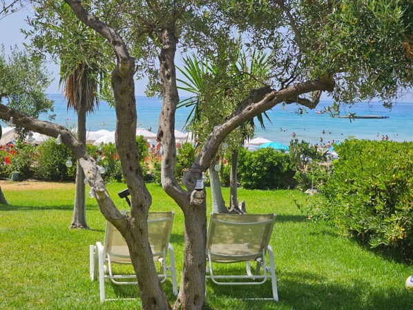 Un espace herbeux avec deux chaises blanches sous un arbre, d’où l’on a une vue sur une plage avec des parasols et des gens.