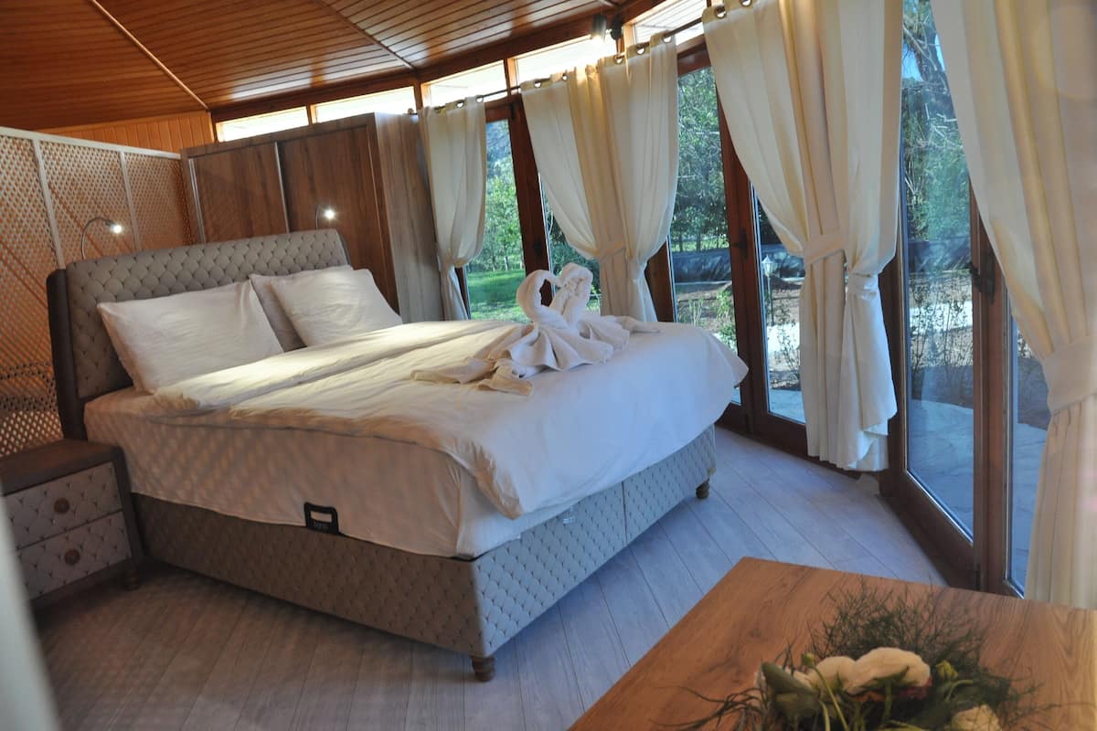 A bedroom with a large bed, a nightstand, a wooden table, and a view of the outdoors through large windows.