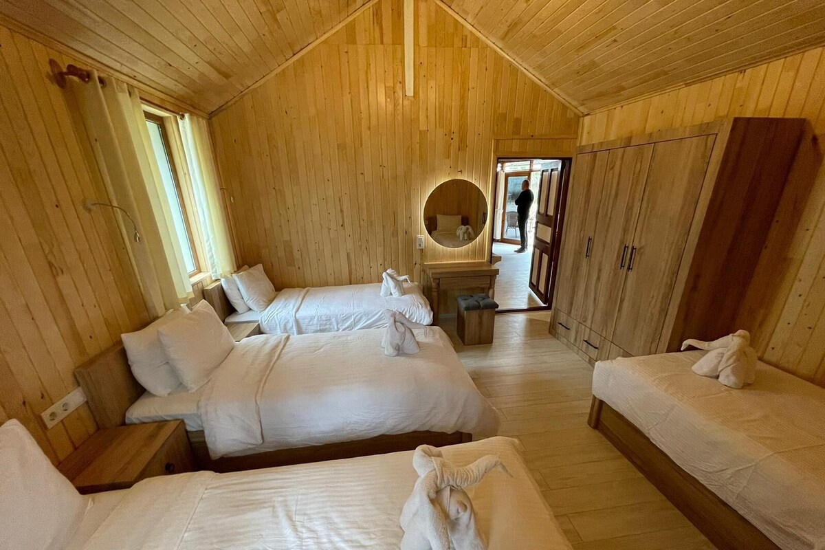 Une pièce aux murs et au plancher en bois, avec deux lits recouverts de draps blancs, un miroir rond et une armoire en bois.