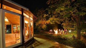 A modern building with large glass doors, a stone pathway, and a fire pit area with people gathered around.