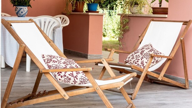 Deux fauteuils de jardin en bois, dotés de coussins blancs et ornés de motifs floraux, sont placés sur une terrasse carrelée. Un mur rose et des plantes en pot se trouvent à l’arrière-plan.