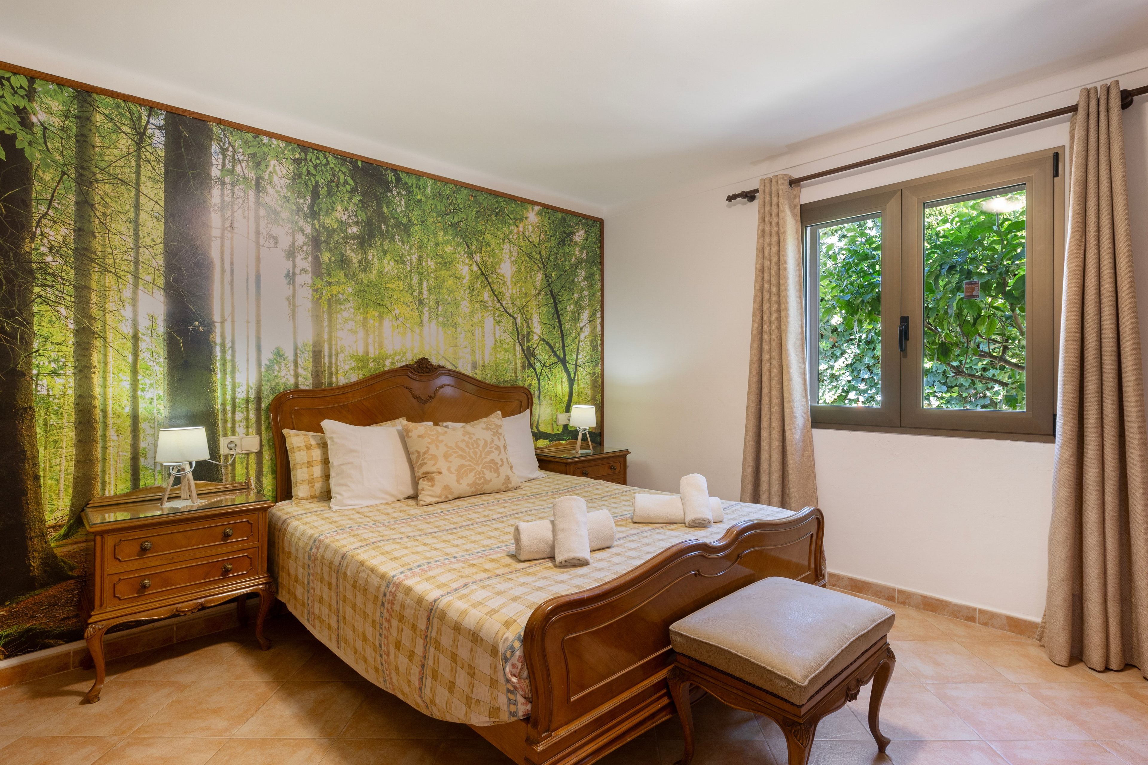 Un dormitorio con un gran mural de un bosque, una cama de madera con un banco, y una ventana con cortinas.