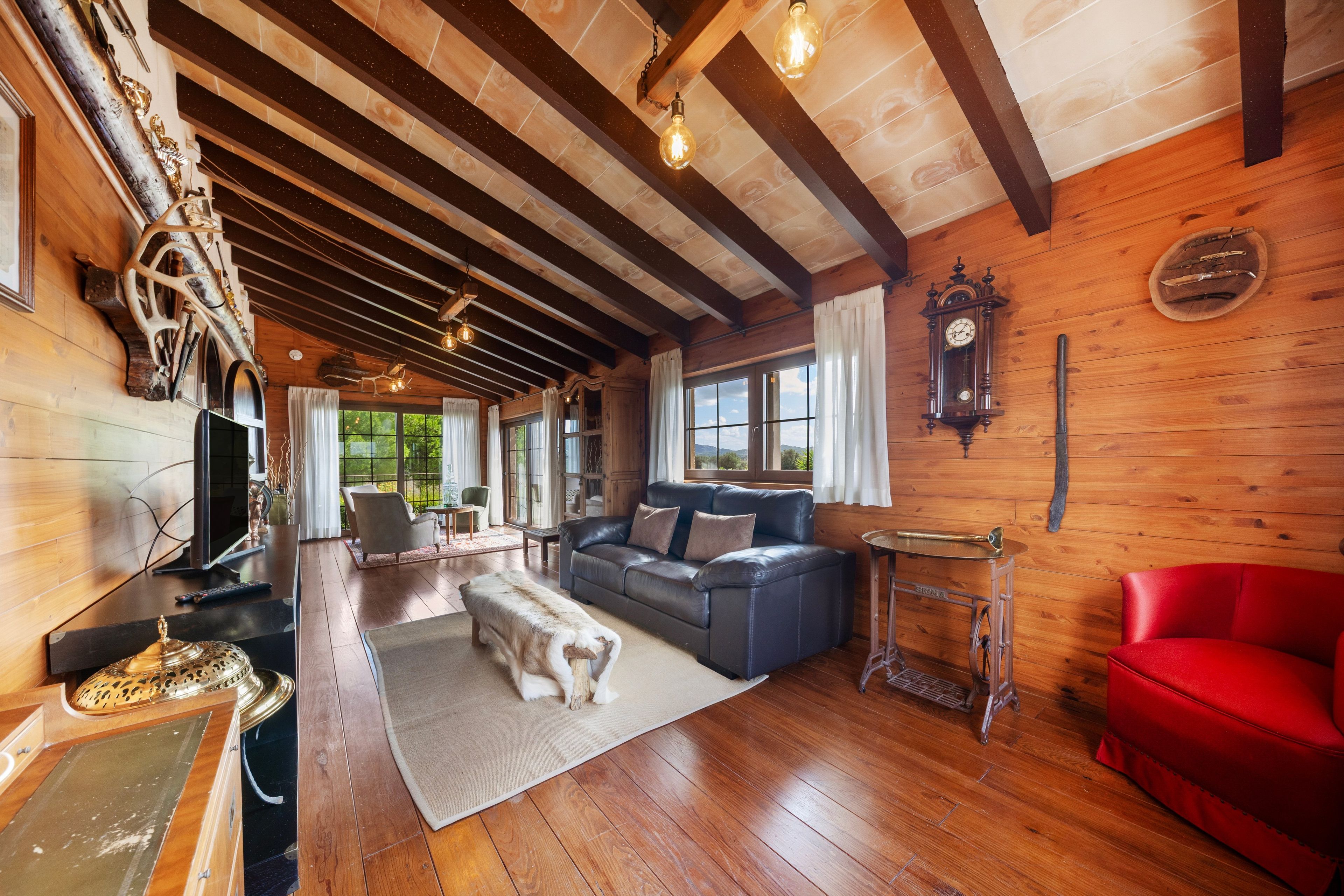 A cozy cabin interior with a wooden ceiling, a sofa, a red chair, a dining table, and a large window offering a view of the outdoors.