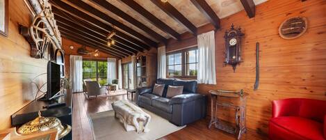 A cozy cabin interior with a wooden ceiling, a sofa, a red chair, a dining table, and a large window offering a view of the outdoors.