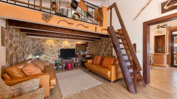 Un espace de vie de style loft avec un plafond haut, des poutres apparentes et un mur en pierre. On y trouve un escalier en bois menant à un niveau supérieur, un canapé en cuir et un téléviseur à écran plat.