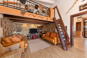 A loft-style living area with a high ceiling, exposed wooden beams, and a stone wall. There is a wooden ladder leading to an upper level, a leather sofa, and a flat-screen TV.