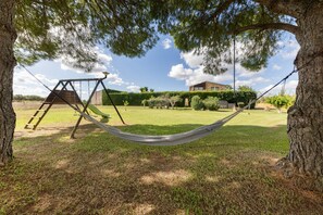 A hammock between trees in a grassy yard with a swing set and a building in the background.