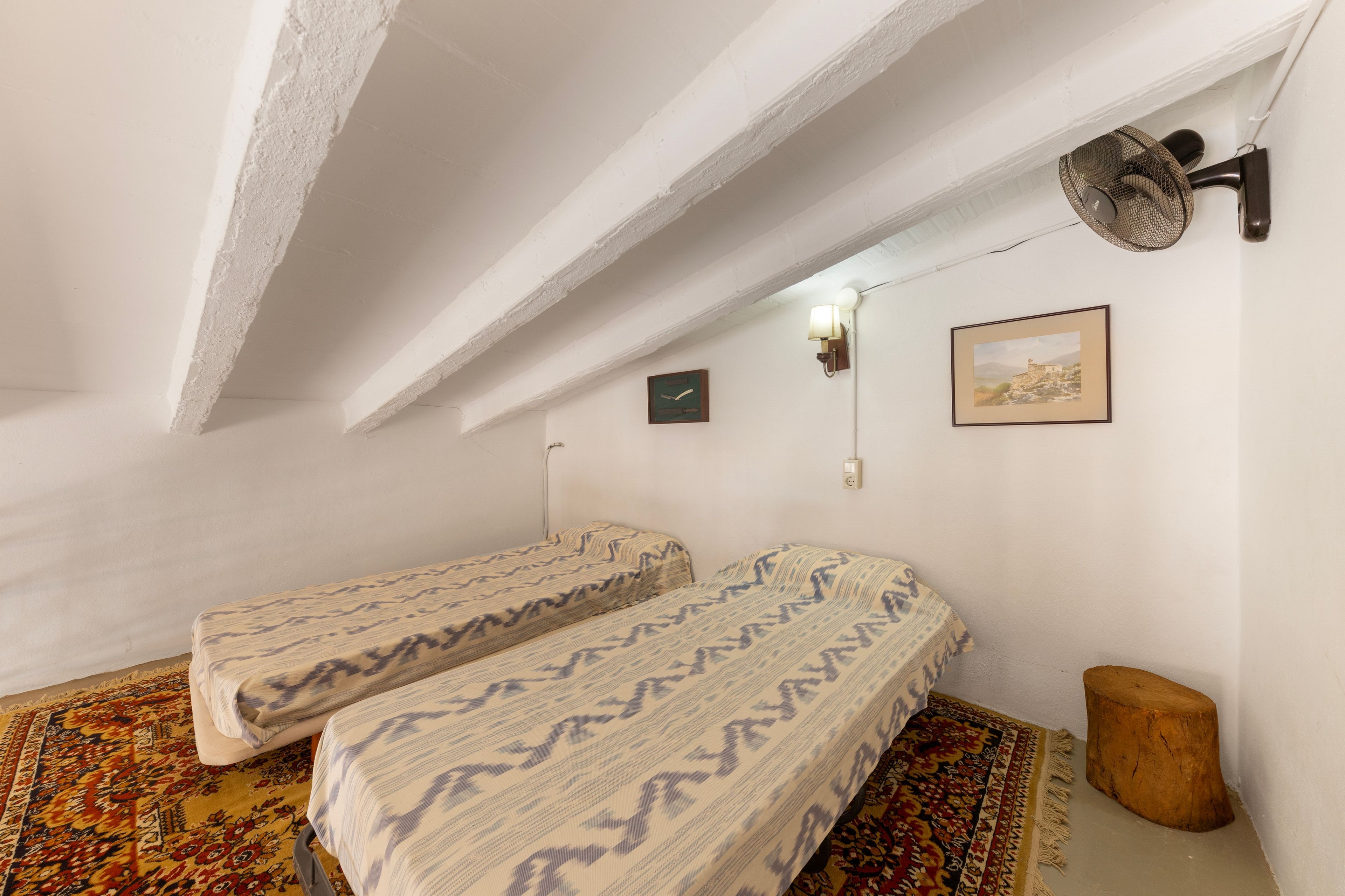 Una habitación con dos camas individuales, un ventilador de pared, una mesita pequeña, una alfombra y un cuadro en la pared.