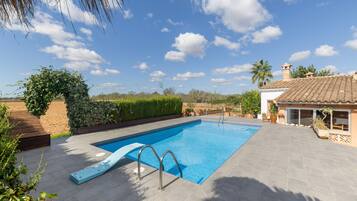 Une piscine rectangulaire avec un toboggan, entourée d’une terrasse agrémentée de plantes en pot et offrant une vue sur le ciel.
